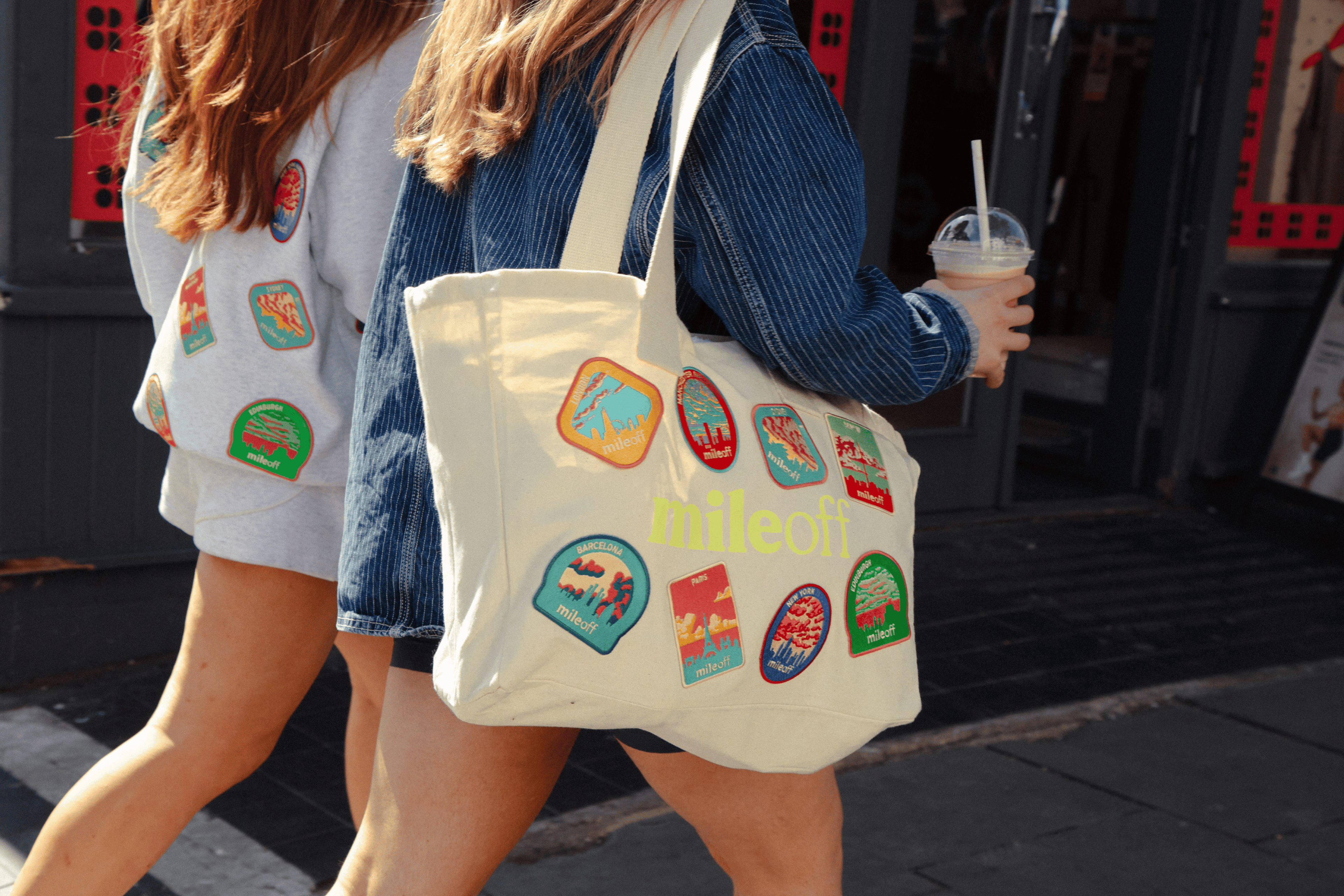 Person holding a tote bag with colorful patches and 'milehoff' text, walking outdoors.
